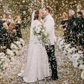 Load image into Gallery viewer, Wedding couple kissing under a shower of white flowers with guests in the background.